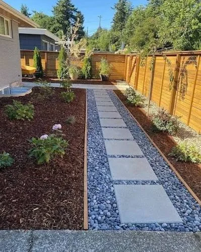 Walkway of square concrete pavers bordered by grey pebble and mulch beds