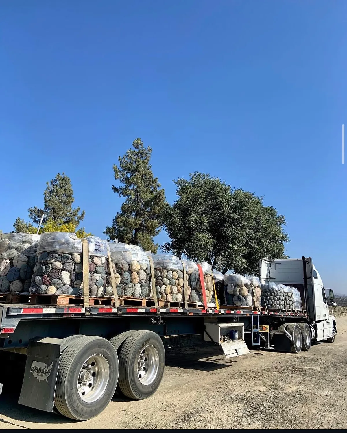 A flatbed truck loaded with strapped wire baskets of beach pebble under a clear blue sky