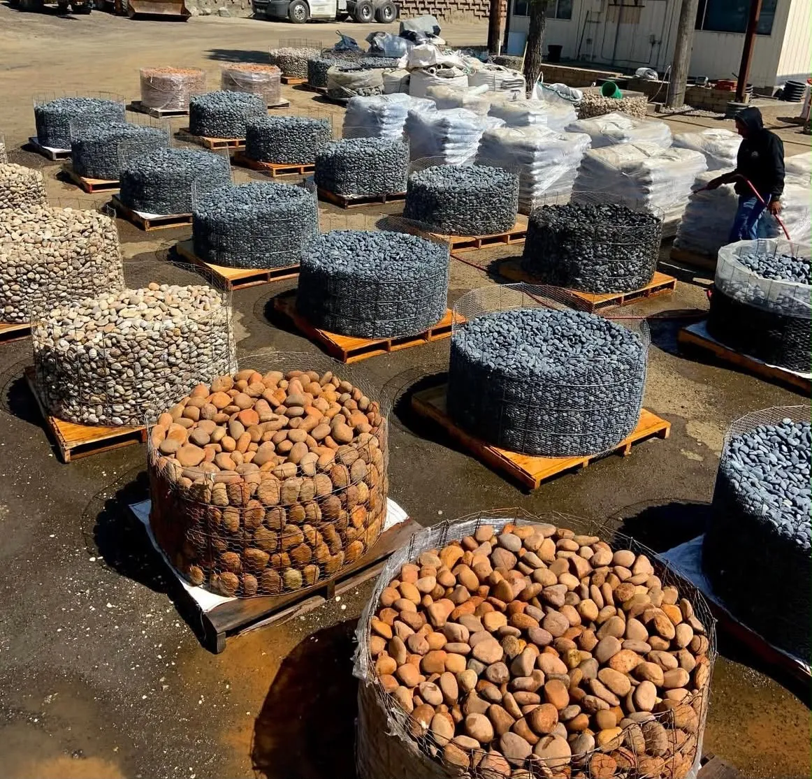 Wire baskets of black, buff and red beach pebble on pallets next to stacks of bagged stone
